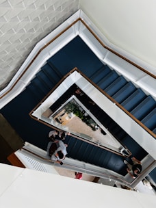 A top-down view of a multi-level staircase with geometric patterns. The stairs are dark blue with white railings and wooden accents. Green plants are visible on one floor and several people are walking or standing on different levels.