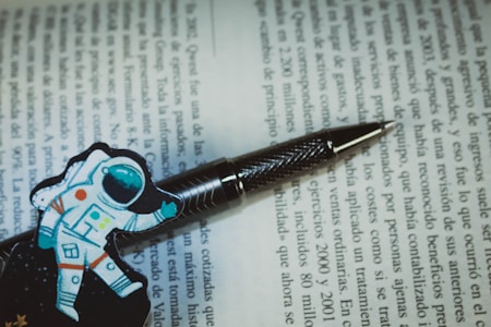 A pen with a textured metallic finish rests on an open book with text in Spanish. Next to the pen, there is a colorful illustration of an astronaut, adding a playful touch to the scholarly setting.