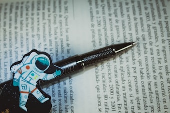 A pen with a textured metallic finish rests on an open book with text in Spanish. Next to the pen, there is a colorful illustration of an astronaut, adding a playful touch to the scholarly setting.