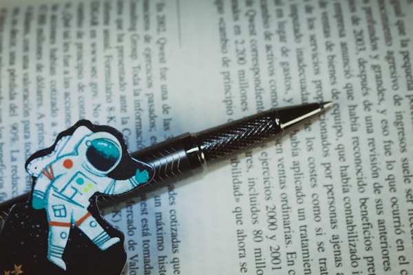A pen with a textured metallic finish rests on an open book with text in Spanish. Next to the pen, there is a colorful illustration of an astronaut, adding a playful touch to the scholarly setting.