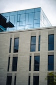 A modern building featuring a combination of stone and glass elements. The lower portion consists of vertical stone panels with tall, narrow windows, while the upper part is made of glass, creating a sleek, reflective surface.