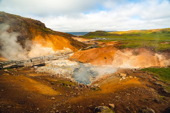 Steam rises from geothermal vents amidst an ochre-colored landscape with rocky terrain. A boardwalk allows for safe viewing, and verdant hills stretch into the distance under a partly cloudy sky.