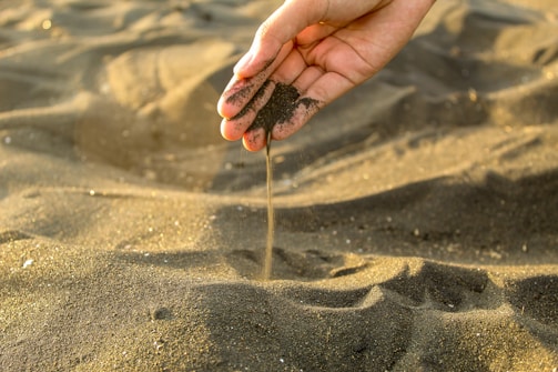 A hand is delicately allowing black sand to flow through the fingers, creating a stream of sand against a backdrop of a beach landscape. The sand appears fine and dark, with a warm light casting subtle highlights and shadows across the scene.