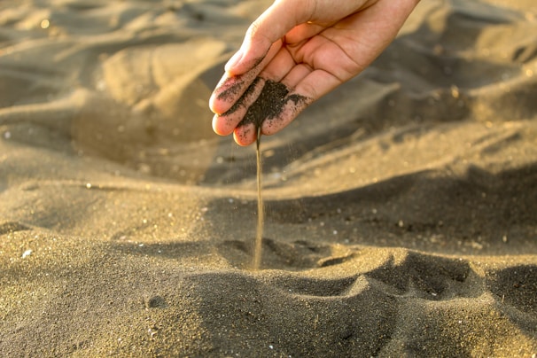 A hand is delicately allowing black sand to flow through the fingers, creating a stream of sand against a backdrop of a beach landscape. The sand appears fine and dark, with a warm light casting subtle highlights and shadows across the scene.