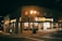 A corner building with a mini mart named Marie's is illuminated by street lamps at night. The structure includes a business sign and other advertisements. It is surrounded by deserted streets and a few parked cars. The building has a brick facade with multiple windows.