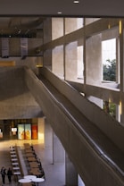 The image depicts the interior of a modern, spacious building featuring large, concrete walls and an indoor ramp. The design includes high ceilings with ample natural light streaming from large windows. There is a seating area with tables and chairs, and two people walking nearby. The overall style is minimalist and industrial.