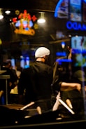 A chef wearing a hairnet stands in a dimly lit kitchen with a focus on cooking utensils in the foreground. Neon signs and bright lights create reflections in the glass, adding vibrant colors to the scene.