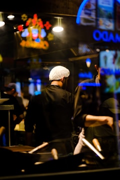 A chef wearing a hairnet stands in a dimly lit kitchen with a focus on cooking utensils in the foreground. Neon signs and bright lights create reflections in the glass, adding vibrant colors to the scene.