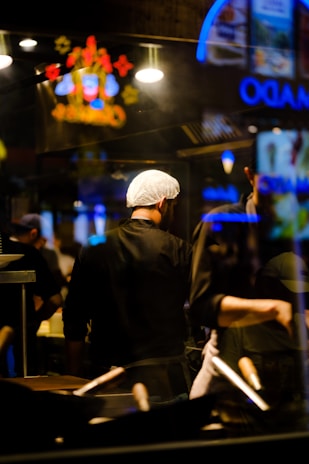 A chef wearing a hairnet stands in a dimly lit kitchen with a focus on cooking utensils in the foreground. Neon signs and bright lights create reflections in the glass, adding vibrant colors to the scene.