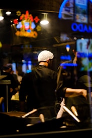 A chef wearing a hairnet stands in a dimly lit kitchen with a focus on cooking utensils in the foreground. Neon signs and bright lights create reflections in the glass, adding vibrant colors to the scene.