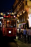 A red tram marked with the destinations 'Taksim' and 'Tunel' is moving along a lively street. The tram is illuminated from within and is surrounded by people walking on both sides. The buildings lining the street are elegantly lit, featuring ornate decorations and glowing lights in the background, indicating a night-time urban setting.