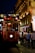 A red tram marked with the destinations 'Taksim' and 'Tunel' is moving along a lively street. The tram is illuminated from within and is surrounded by people walking on both sides. The buildings lining the street are elegantly lit, featuring ornate decorations and glowing lights in the background, indicating a night-time urban setting.