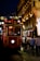 A red tram marked with the destinations 'Taksim' and 'Tunel' is moving along a lively street. The tram is illuminated from within and is surrounded by people walking on both sides. The buildings lining the street are elegantly lit, featuring ornate decorations and glowing lights in the background, indicating a night-time urban setting.