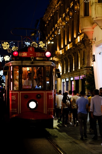 A red tram marked with the destinations 'Taksim' and 'Tunel' is moving along a lively street. The tram is illuminated from within and is surrounded by people walking on both sides. The buildings lining the street are elegantly lit, featuring ornate decorations and glowing lights in the background, indicating a night-time urban setting.