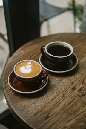 Two cups of coffee sit on a wooden table in a cafe setting. One cup features a latte with a leaf-shaped latte art design, while the other cup contains black coffee. The overall atmosphere is cozy and inviting.