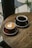 Two cups of coffee sit on a wooden table in a cafe setting. One cup features a latte with a leaf-shaped latte art design, while the other cup contains black coffee. The overall atmosphere is cozy and inviting.