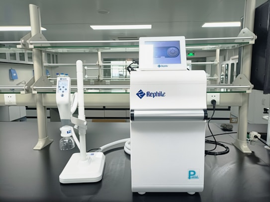 A laboratory setting featuring a water purification system with the brand name Rephile displayed on it. The setup includes a control unit with a digital display screen and an attached dispenser with a control panel. The background shows lab shelves and equipment, indicating a clean, sterile environment.