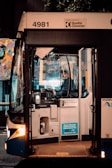 A bus driver is seated inside a bus, focused on driving or preparing to drive. The front door of the bus is open, and there is a sticker indicating payment options on the outside. The surroundings are dimly lit, suggesting it is either early morning or late evening. A colorful mural can be seen in the background through the bus windows. The bus has the number 4981 and is operated by Keolis Downer.