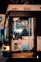 A bus driver is seated inside a bus, focused on driving or preparing to drive. The front door of the bus is open, and there is a sticker indicating payment options on the outside. The surroundings are dimly lit, suggesting it is either early morning or late evening. A colorful mural can be seen in the background through the bus windows. The bus has the number 4981 and is operated by Keolis Downer.