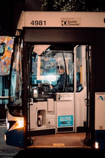 A bus driver is seated inside a bus, focused on driving or preparing to drive. The front door of the bus is open, and there is a sticker indicating payment options on the outside. The surroundings are dimly lit, suggesting it is either early morning or late evening. A colorful mural can be seen in the background through the bus windows. The bus has the number 4981 and is operated by Keolis Downer.