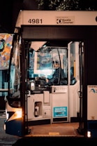 A bus driver is seated inside a bus, focused on driving or preparing to drive. The front door of the bus is open, and there is a sticker indicating payment options on the outside. The surroundings are dimly lit, suggesting it is either early morning or late evening. A colorful mural can be seen in the background through the bus windows. The bus has the number 4981 and is operated by Keolis Downer.