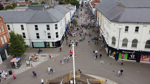A busy urban street with people walking and shopping. The view is from above, showcasing a pedestrian area surrounded by shops and buildings. A Union Jack flag is visible in the foreground, and there are various storefronts, including Next and Superdrug. Floral decorations and a telephone booth are present on the left side.