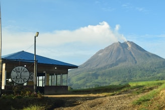 A small coffee shop with a blue roof and large windows is situated in the foreground. The backdrop features a picturesque mountain emitting some smoke or mist, surrounded by lush green vegetation under a clear blue sky.