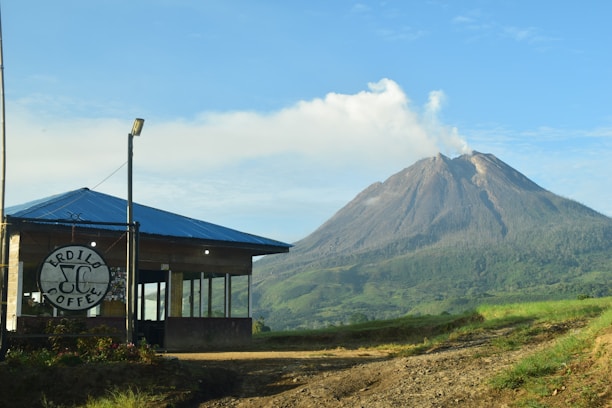 A small coffee shop with a blue roof and large windows is situated in the foreground. The backdrop features a picturesque mountain emitting some smoke or mist, surrounded by lush green vegetation under a clear blue sky.
