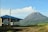 A small coffee shop with a blue roof and large windows is situated in the foreground. The backdrop features a picturesque mountain emitting some smoke or mist, surrounded by lush green vegetation under a clear blue sky.