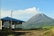 A small coffee shop with a blue roof and large windows is situated in the foreground. The backdrop features a picturesque mountain emitting some smoke or mist, surrounded by lush green vegetation under a clear blue sky.