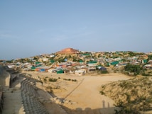 A sprawling settlement on a hill, featuring numerous makeshift homes constructed from a variety of materials. The homes are arranged in a somewhat organized manner, with some patches of greenery visible between them. A large building with a red roof dominates the hilltop. The foreground shows a sandy area with a series of arranged, circular concrete forms, possibly erosion control or a pathway.