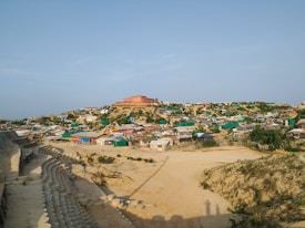 A sprawling settlement on a hill, featuring numerous makeshift homes constructed from a variety of materials. The homes are arranged in a somewhat organized manner, with some patches of greenery visible between them. A large building with a red roof dominates the hilltop. The foreground shows a sandy area with a series of arranged, circular concrete forms, possibly erosion control or a pathway.