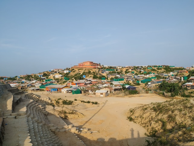 A sprawling settlement on a hill, featuring numerous makeshift homes constructed from a variety of materials. The homes are arranged in a somewhat organized manner, with some patches of greenery visible between them. A large building with a red roof dominates the hilltop. The foreground shows a sandy area with a series of arranged, circular concrete forms, possibly erosion control or a pathway.