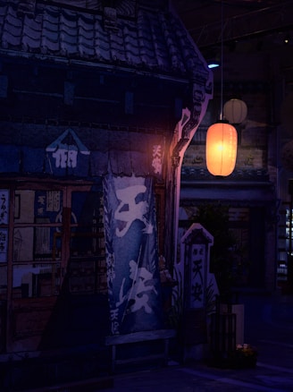 A traditional Japanese storefront is illuminated by the soft glow of an orange paper lantern, casting warm light onto the dark, rustic exterior. The building features detailed architectural elements with wooden doors and panels, and Japanese characters are visible on banners and signs.