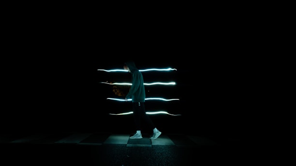 A person in a hoodie is walking across a dark space with streaks of light trailing behind them, creating a sense of motion and mystery.