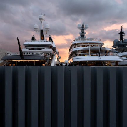 Two large luxury yachts are docked side by side, partially obscured by a dark, corrugated fence in the foreground. The yachts are equipped with multiple decks, antennas, and observation equipment on their upper levels. The sky in the background is a dramatic mix of dark clouds and warm hues of pink and orange, indicating a sunset or sunrise.