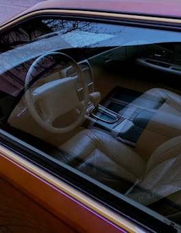 The interior of a car is visible through the window, showcasing a steering wheel, dashboard, and leather seats. The seats and interior elements appear in neutral tones, and there's a reflection of outdoor trees on the window.