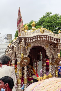 A decorated chariot adorned with intricate metallic carvings and golden ornaments is surrounded by a group of people. The chariot features traditional motifs and is topped with a colorful flag. The scene appears to be part of a cultural or religious procession.