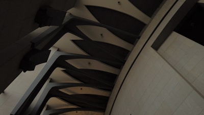 A series of large, dark structural beams and columns feature prominently, casting shadows and creating a pattern of converging lines and smooth curves on a textured stone wall. The overall design gives a sense of architectural depth and complexity.