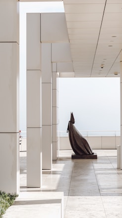 A modern architectural space features tall, white columns and a sleek, minimal design with clean lines. The setting appears to be an outdoor or semi-outdoor area, open to natural light. A large, abstract sculpture stands prominently in the middle of the walkway, casting soft shadows on the tiled floor.