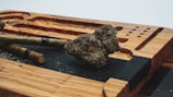 A wooden tray with carved grooves holds two cigar-like objects and a large, dense nugget of cannabis. The tray has a natural wood grain finish and features an engraved pattern. The surface is relatively clean, with scattered crumbs of material.