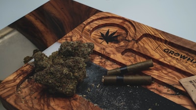 A wooden tray with a carved cannabis leaf design holds cannabis buds and two rolled blunts. The tray is engraved with the name 'GrowHub'. A small part of a brown paper bag is visible on the side.