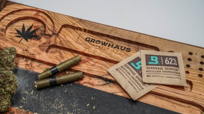 A wooden surface with a carved cannabis leaf and the word 'Growhaus'. There are several cannabis joints and buds next to two terpene shield humidity packets.