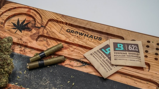 A wooden surface with a carved cannabis leaf and the word 'Growhaus'. There are several cannabis joints and buds next to two terpene shield humidity packets.