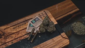A wooden cutting board with a branded logo is used as a surface for displaying cannabis products, including several large buds and a pile of ground herb. There are also rolling papers or pre-rolled cones and humidity control packs on the board.