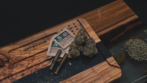 A wooden table holds several items related to cannabis consumption, including buds of cannabis, packaging materials, two pre-rolled joints, and a pile of ground cannabis. The table also has an engraved design.