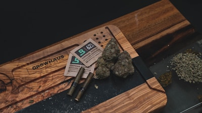 A wooden table holds several items related to cannabis consumption, including buds of cannabis, packaging materials, two pre-rolled joints, and a pile of ground cannabis. The table also has an engraved design.