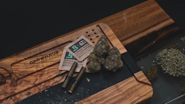 A wooden table holds several items related to cannabis consumption, including buds of cannabis, packaging materials, two pre-rolled joints, and a pile of ground cannabis. The table also has an engraved design.