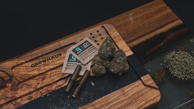 A wooden table holds several items related to cannabis consumption, including buds of cannabis, packaging materials, two pre-rolled joints, and a pile of ground cannabis. The table also has an engraved design.