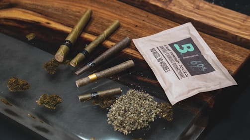 Several rolled cigars or blunts are placed on a wooden surface next to a humidity control packet. There is a display of scattered dried herbs or leaves.